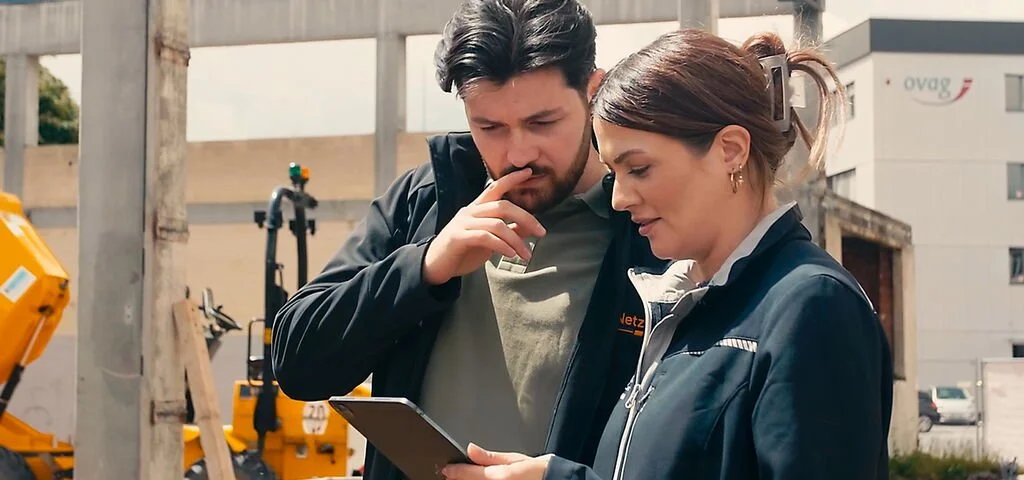 Zwei Menschen diskutieren auf einer Baustelle, schauen auf ein Tablet, im Hintergrund Baustellengeräte und Gebäude.