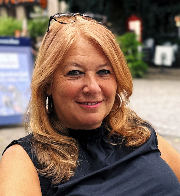 Porträt einer lächelnden roten-haired Frau mit Sonnenbrille auf dem Kopf, in einem schwarzen Oberteil, sitzt im Freien auf einer gepflasterten Fläche, im Hintergrund Bäume und Hinweise auf eine Stadtumgebung.