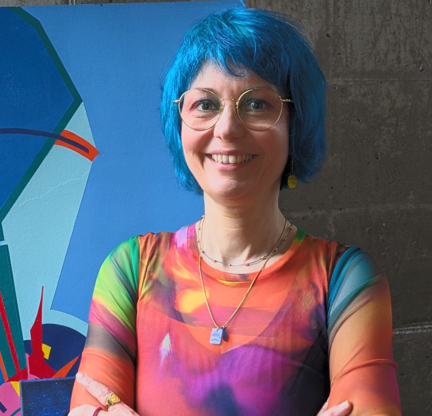Eine Frau mit blauer Kurzhaarfrisur, Brille und buntem, auffälligem Kleid, stehend vor einer Wand und einem farbenfrohen Kunstwerk.