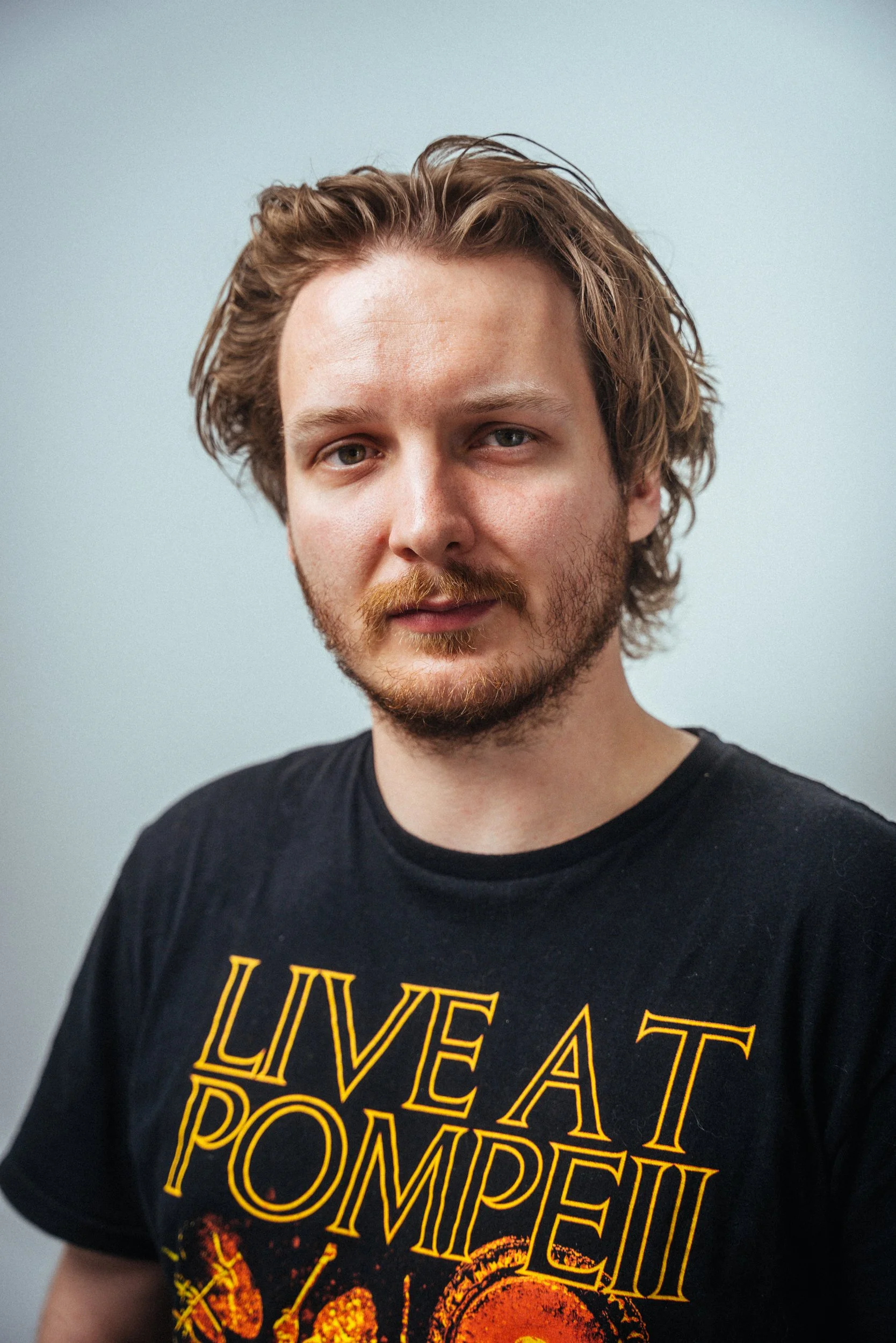 Porträt eines jungen Mannes mit braunem, lockigem Haar und einem Bart, trägt ein schwarzes T-Shirt mit gelber Schrift und orangenem Design, vor hellem, neutralem Hintergrund.