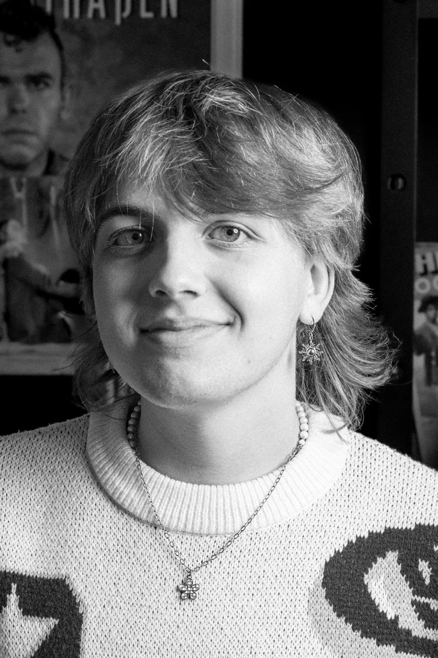 Junge Frau mit Kurzhaarfrisur trägt Ohrringe, zwei Halsketten und ein gestricktes Oberteil, im Hintergrund Poster