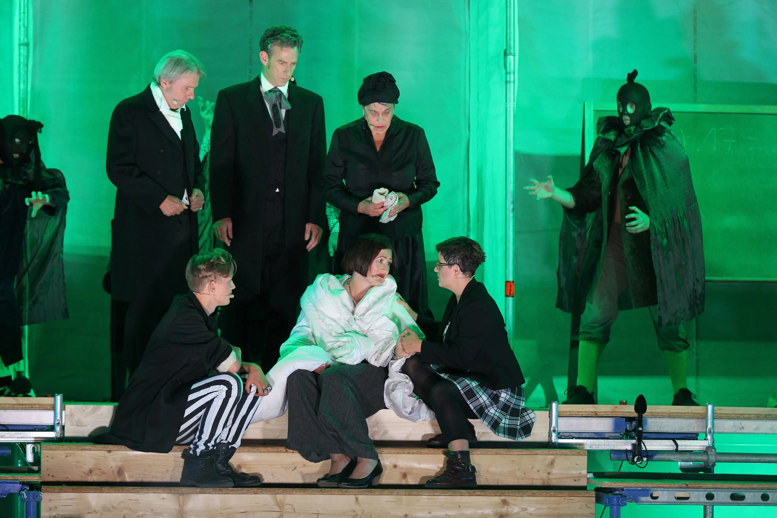 Theaterstück mit mehreren Darstellern, aufwendige Kostüme, düstere Beleuchtung in grün, Szene mit einer Frau in weißen Kleid sitzt auf einer Bank, umgeben von jüngeren Frauen in schwarzen Outfits, zwei Männer stehen neben ihr, eine Person in dunkler 