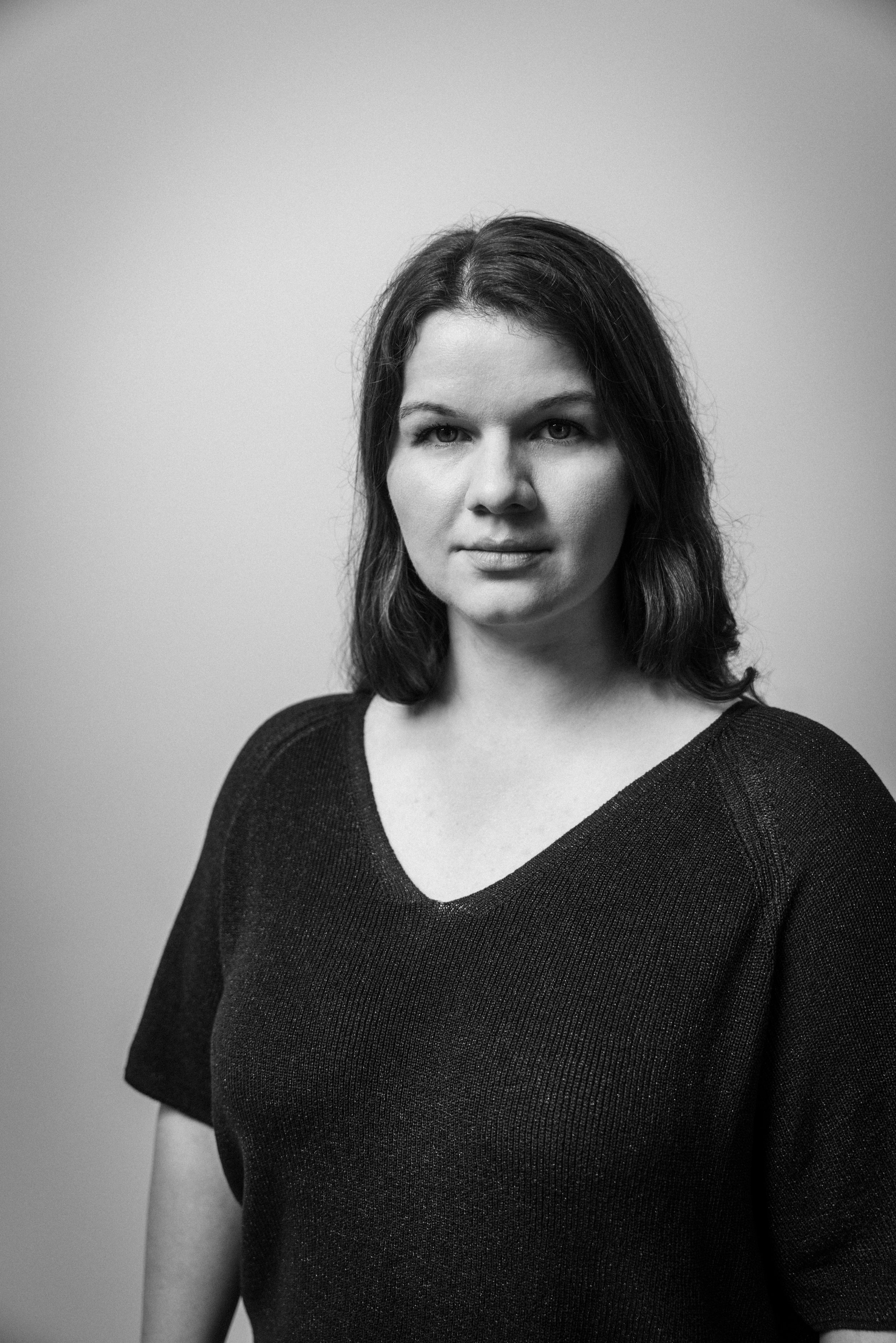 Schwarzweißportrait einer jungen Frau mit dunklem, schulterlangem Haar, die ein schwarzes Oberteil trägt, vor hellem Hintergrund.