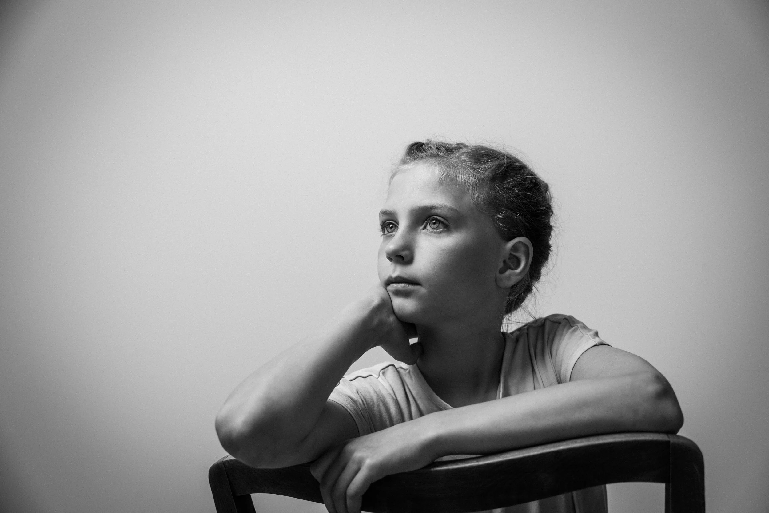 Schwarz-Weiß-Fotografie eines jungen Mädchens, das auf einem Stuhl sitzt, den Kopf auf die Hand stützt und nach oben blickt, vor einem neutralen Hintergrund.