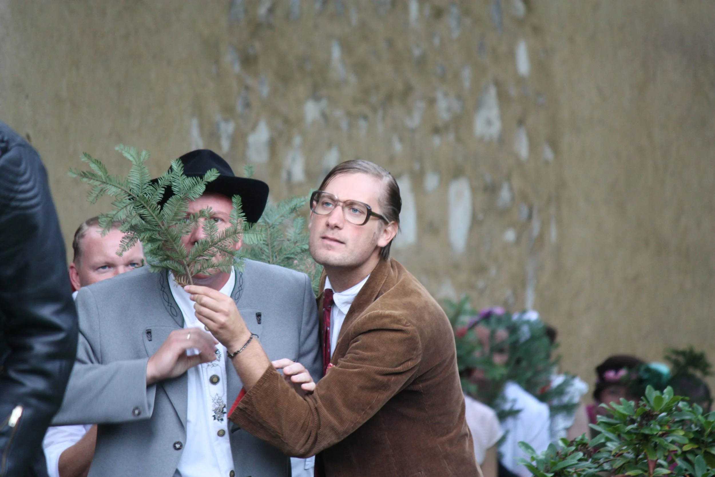 Menschen in traditionellen deutschen Trachten bei einer Veranstaltung, einer hält ein Tannenzweigholz, im Hintergrund weitere Personen mit Blumentöpfen.