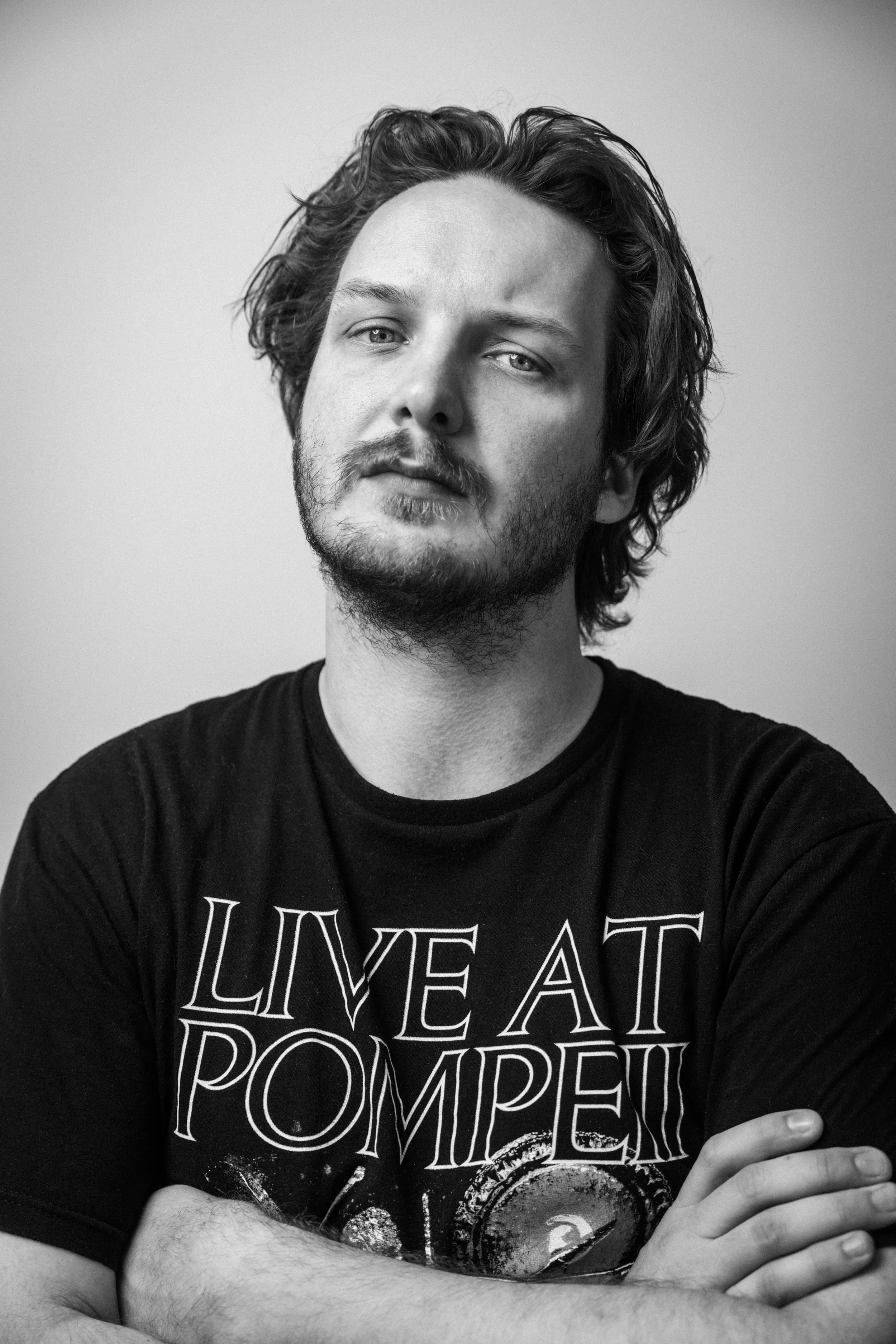 Schwarz-weiß-Fotografie eines Mannes mit langen, gewellten Haaren, leichtem Bart, der eine schwarze T-Shirt trägt. Der Blick ist direkt in die Kamera, die Arme sind vor der Brust verschränkt.