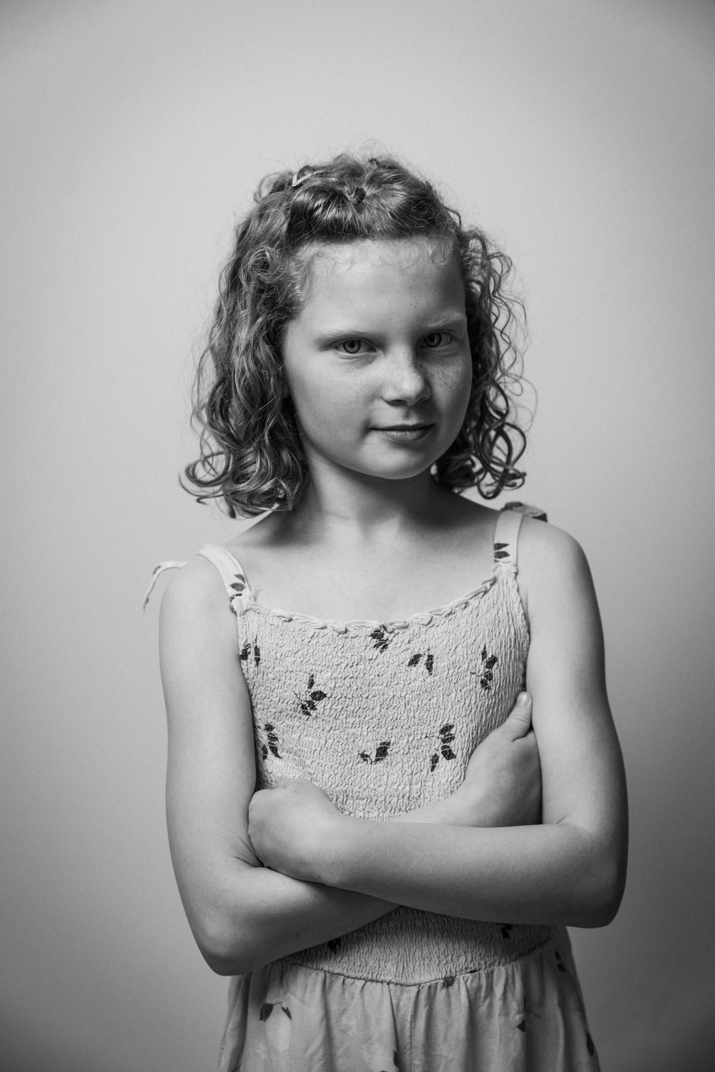 Schwarz-weiß Portrait eines jungen Mädchens mit lockigem Haar, das die Arme verschränkt trägt ein ärmelloses, gemustertes Kleid.