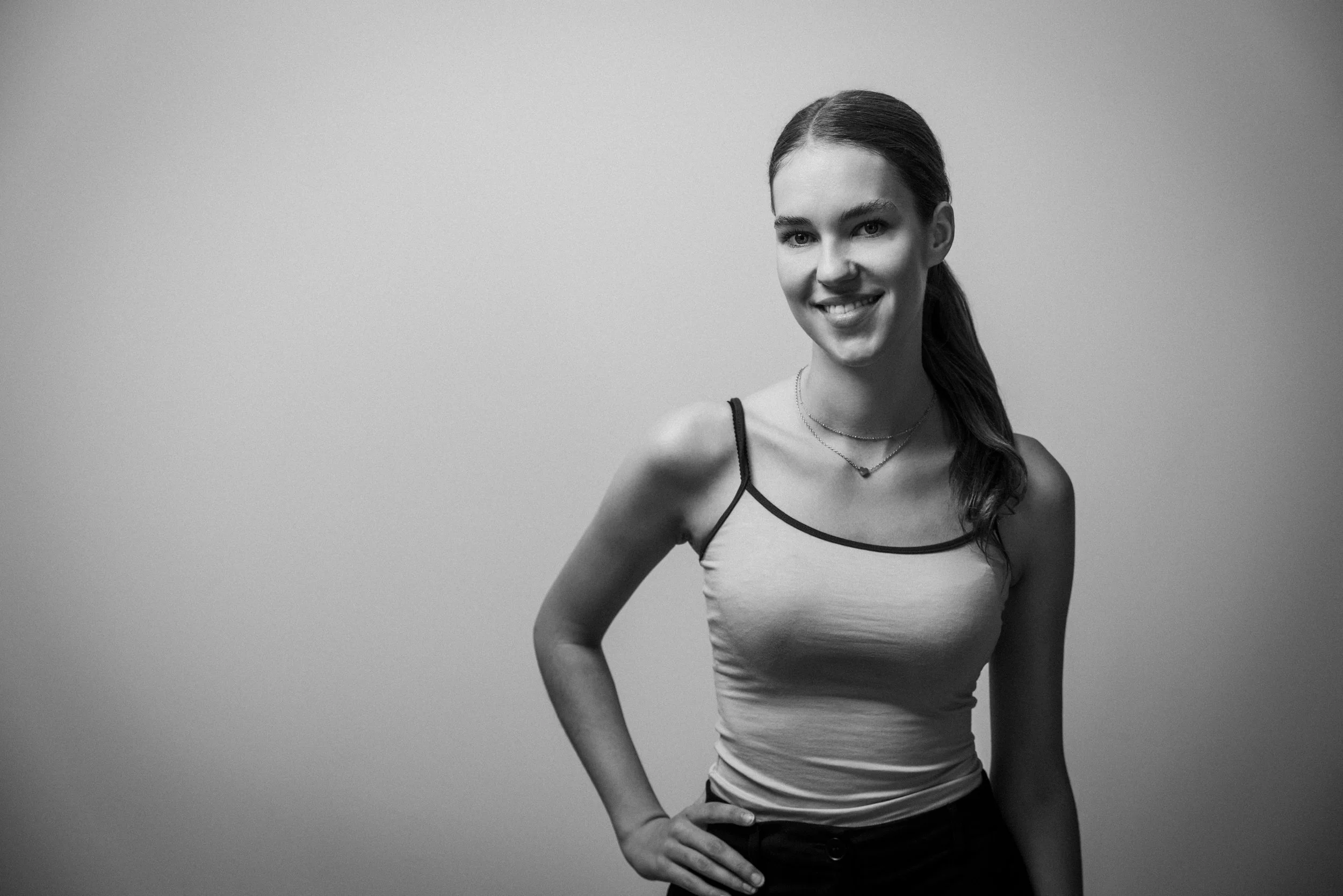 Schwarz-weiß Foto einer jungen Frau mit dunklem langem Haar, die lächelnd vor einem schlichten Hintergrund steht, mit einer Hand auf der Taille, getragen in einem sommerlichen Top mit dünnen Trägern.