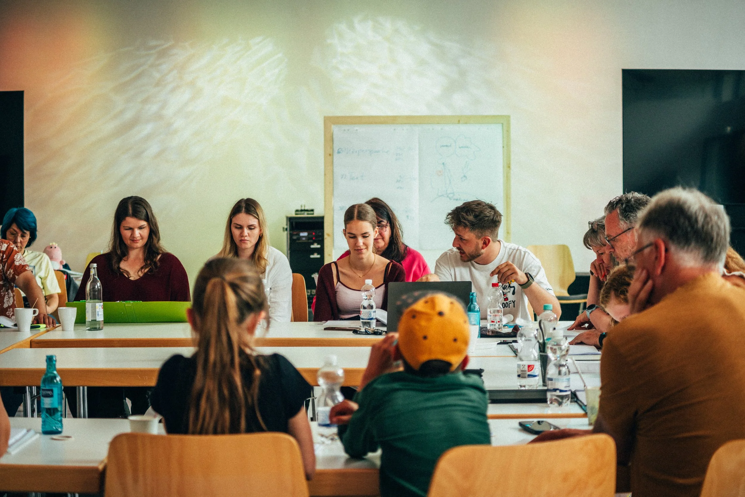 Menschen sitzen um Tische in einem Raum, scheinbar in einem Meeting oder Workshop, mit einem Whiteboard im Hintergrund.