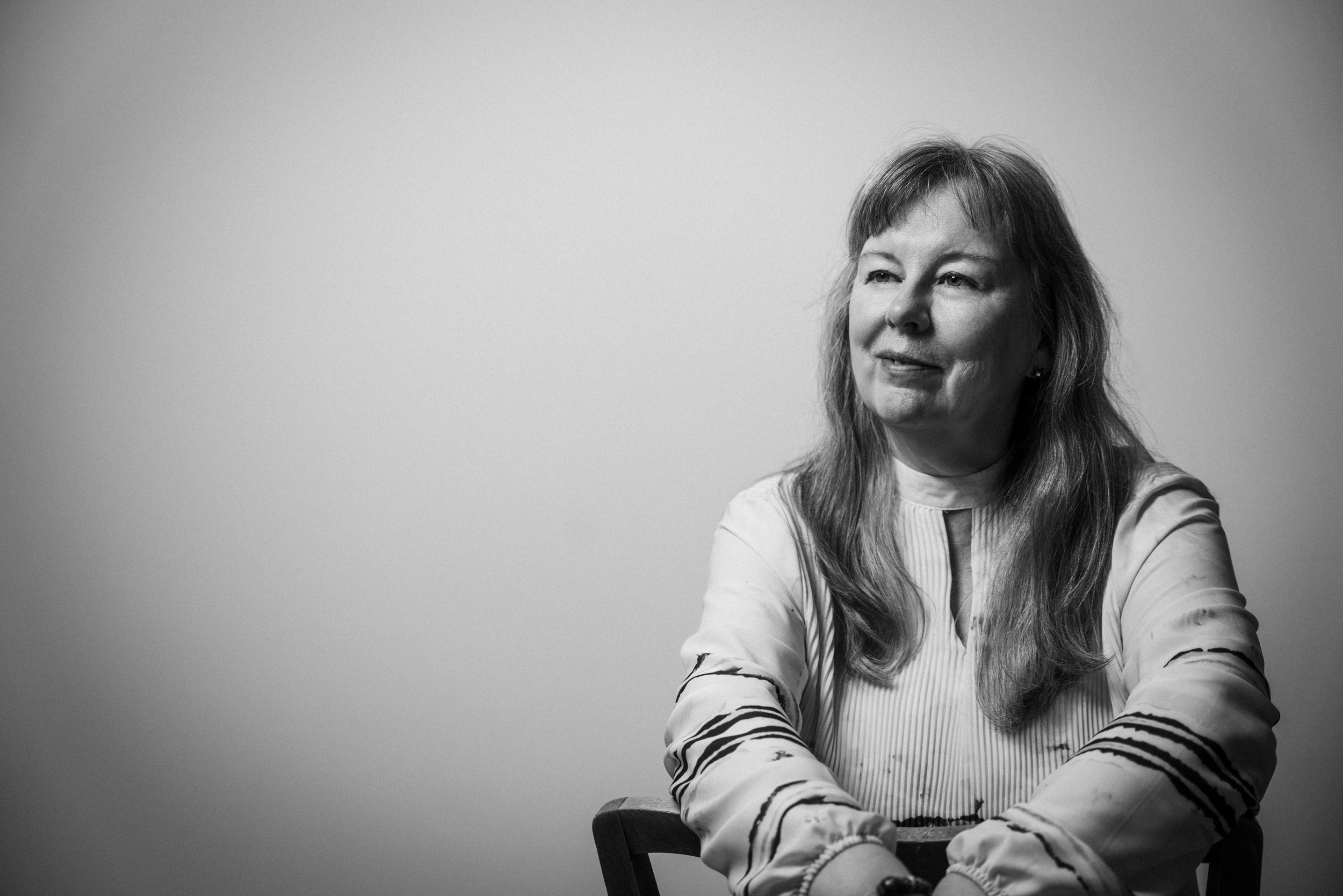 Schwarz-weiß-Foto einer lächelnden Frau mit mittellangem, glattem Haar, die auf einem Stuhl sitzt und den Arm auf den Stuhl lehnt, vor einem schlichten weißen Hintergrund.