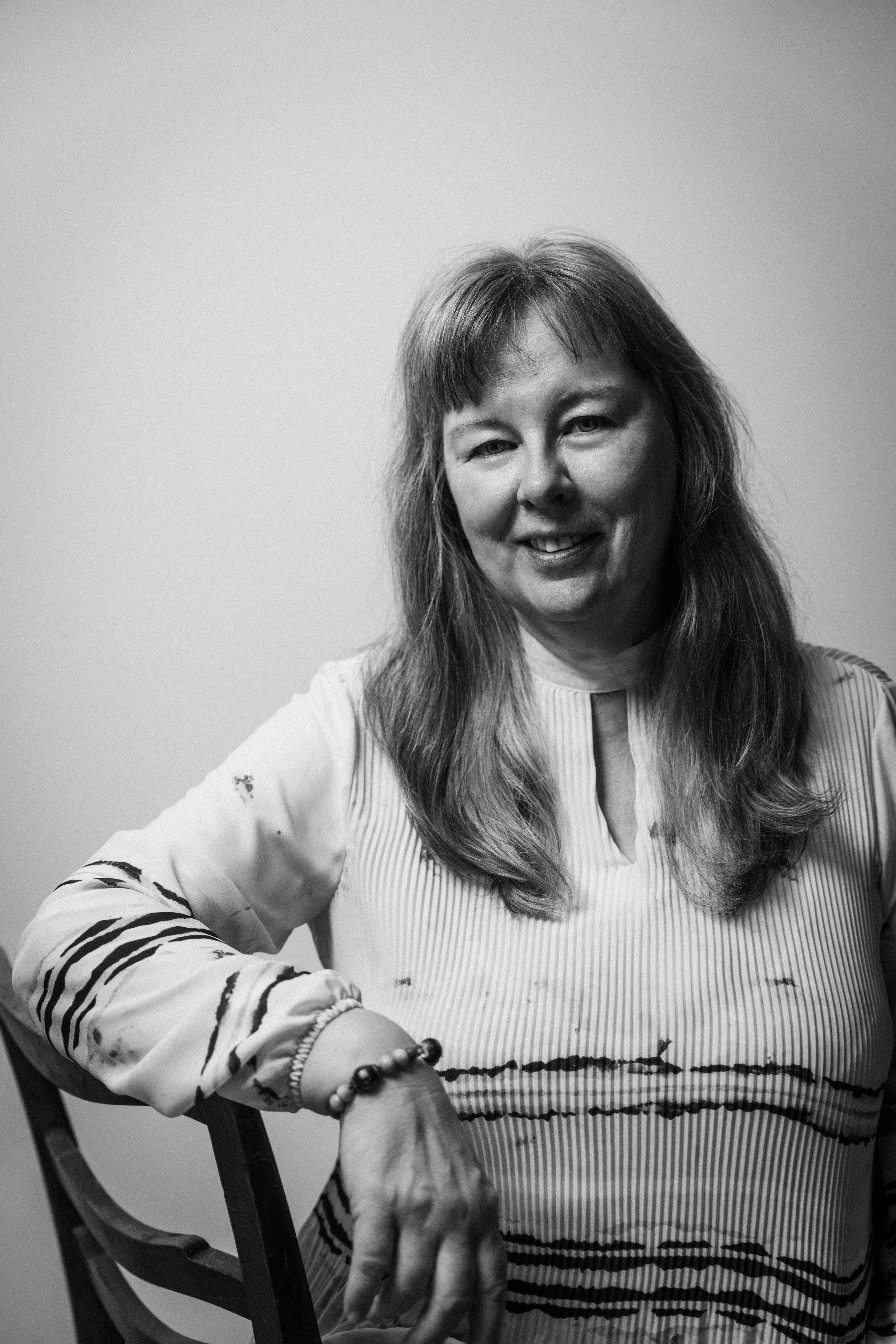Schwarz-Weiß-Bild einer lachenden Frau mit langem Haar, die auf einem Stuhl sitzt und in die Kamera schaut. Sie trägt ein gestreiftes Oberteil. Beate Moritz-Mayer  in  Das Wilde Weib – Das Musical · Birsteiner Festspiele 2027
