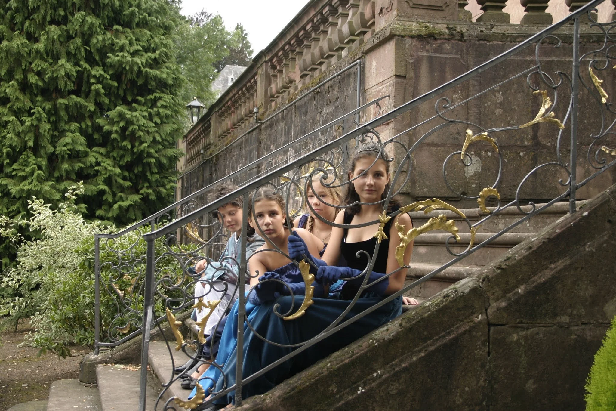 Vier Kinder in Kostümen sitzen auf Steinstufen hinter einem schmiedeeisernen Geländer vor einem historischen Gebäude, umgeben von grünen Bäumen.