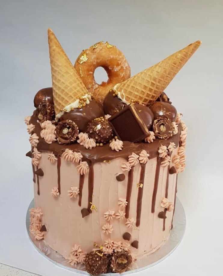 Chocolate frosted cake decorated with ice cream cones, a donut, chocolates, and gold embellishments.