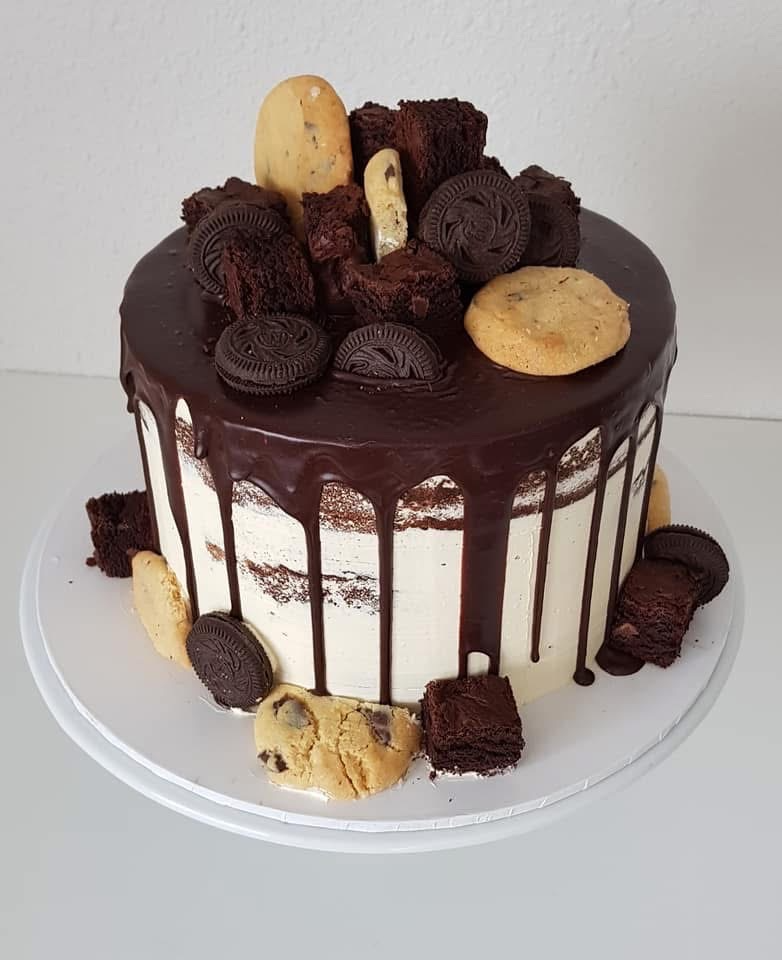 Chocolate drip cake topped with chocolate and vanilla cookies, brownie pieces, and mini cookies on a white plate.