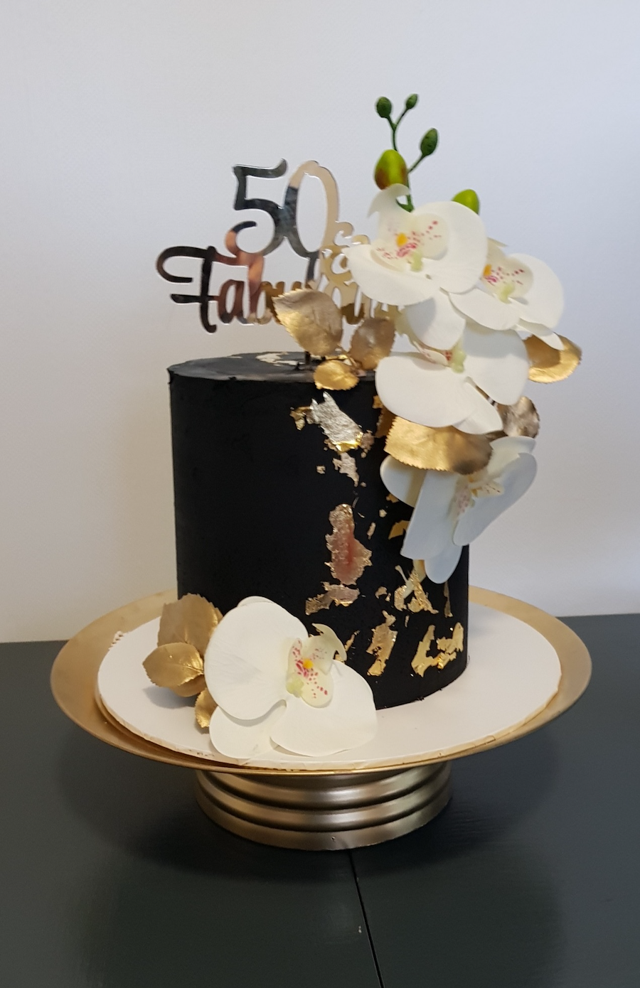 Black anniversary cake decorated with white orchids, gold accents, and a "50th" cake topper.