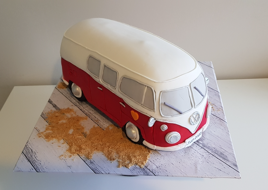 A cake shaped like a red and white vintage Volkswagen camper van on a wooden surface with crumbs around it.
