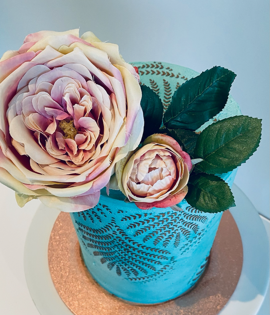 A cake decorated with blue piping and a pattern of black leaves, topped with three large pink and cream roses and green leaves, placed on a white and copper-colored round base.