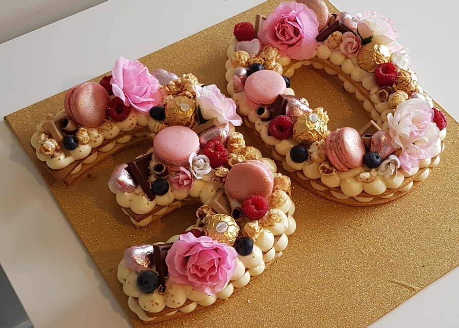 A number two shaped cake decorated with pink flowers, macarons, berries, chocolate pieces, and gold accents, placed on a gold surface.