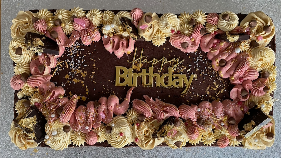 Rectangular birthday cake with chocolate, pink and cream frosting decorations, chocolate and gold accents, and a 'Happy Birthday' gold message in the center.