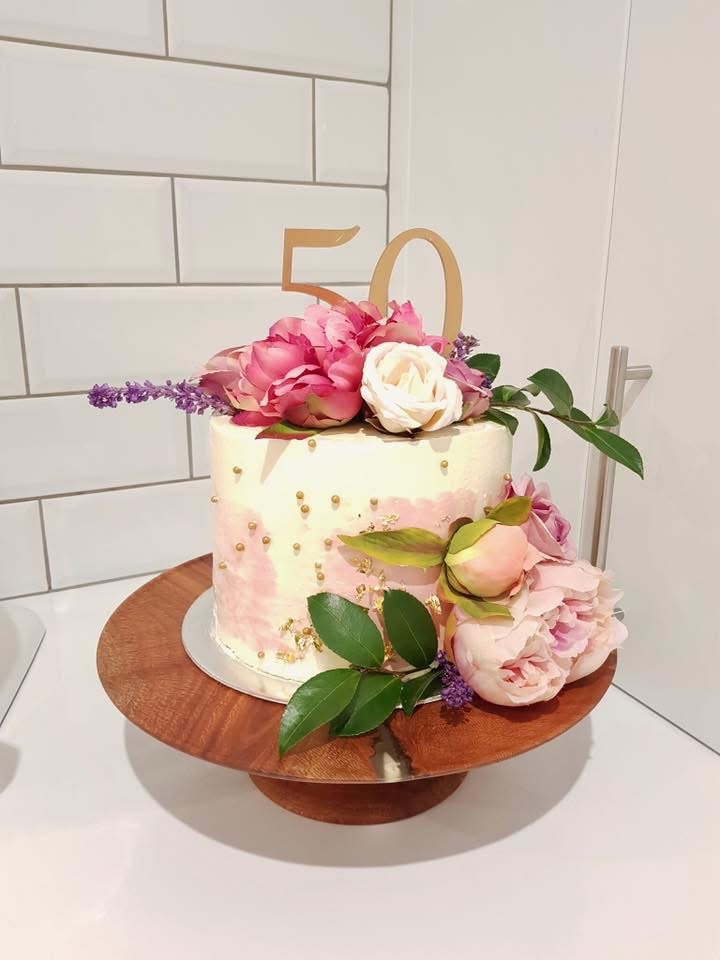 A white birthday cake decorated with pink and white flowers, gold accents, and a '50' topper, placed on a wooden cake stand.
