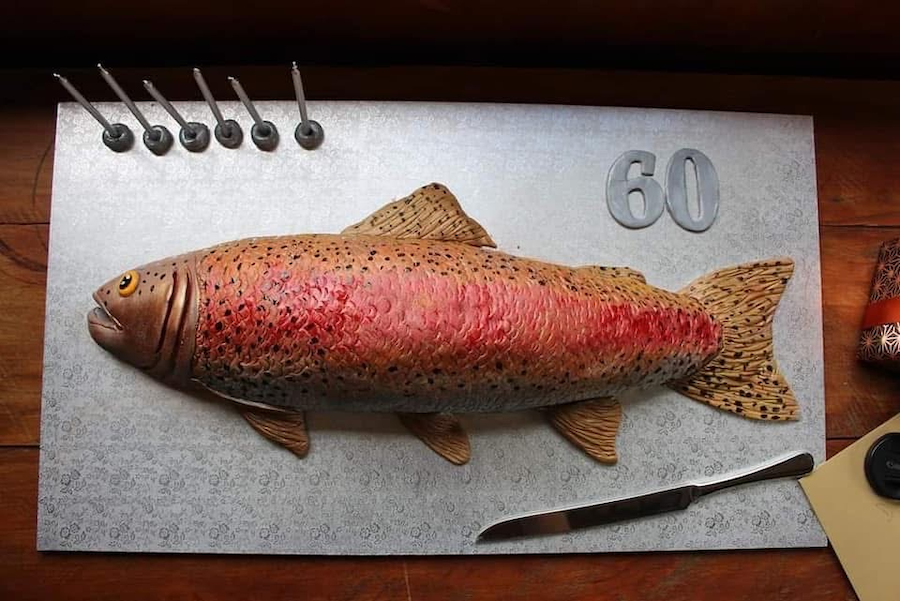 A realistic fish-shaped cake with colorful icing, placed on a decorative surface. The cake is accompanied by a small knife, six nails at the top, and a silver number '60' sign, indicating it might be for a birthday celebration.