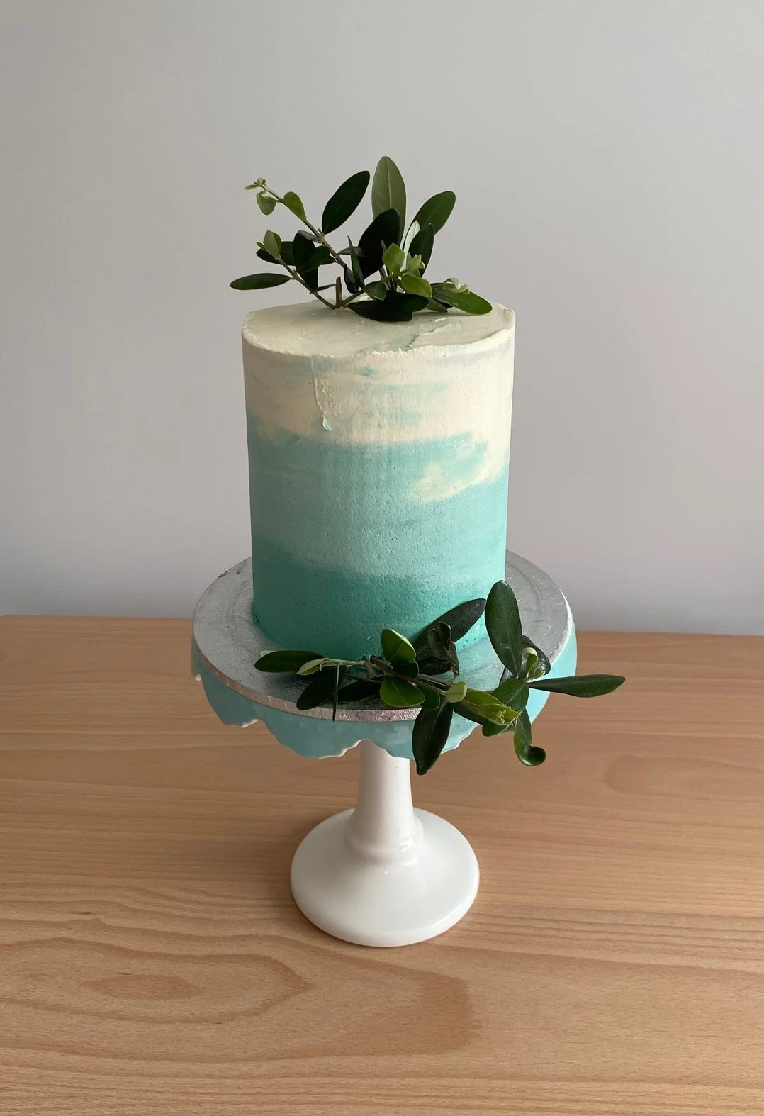 A small gluten free lemon, coconut and almond cake with vanilla buttercream in an ombre style coloured with 'tiffany blue', olive tree leaves to decorate.