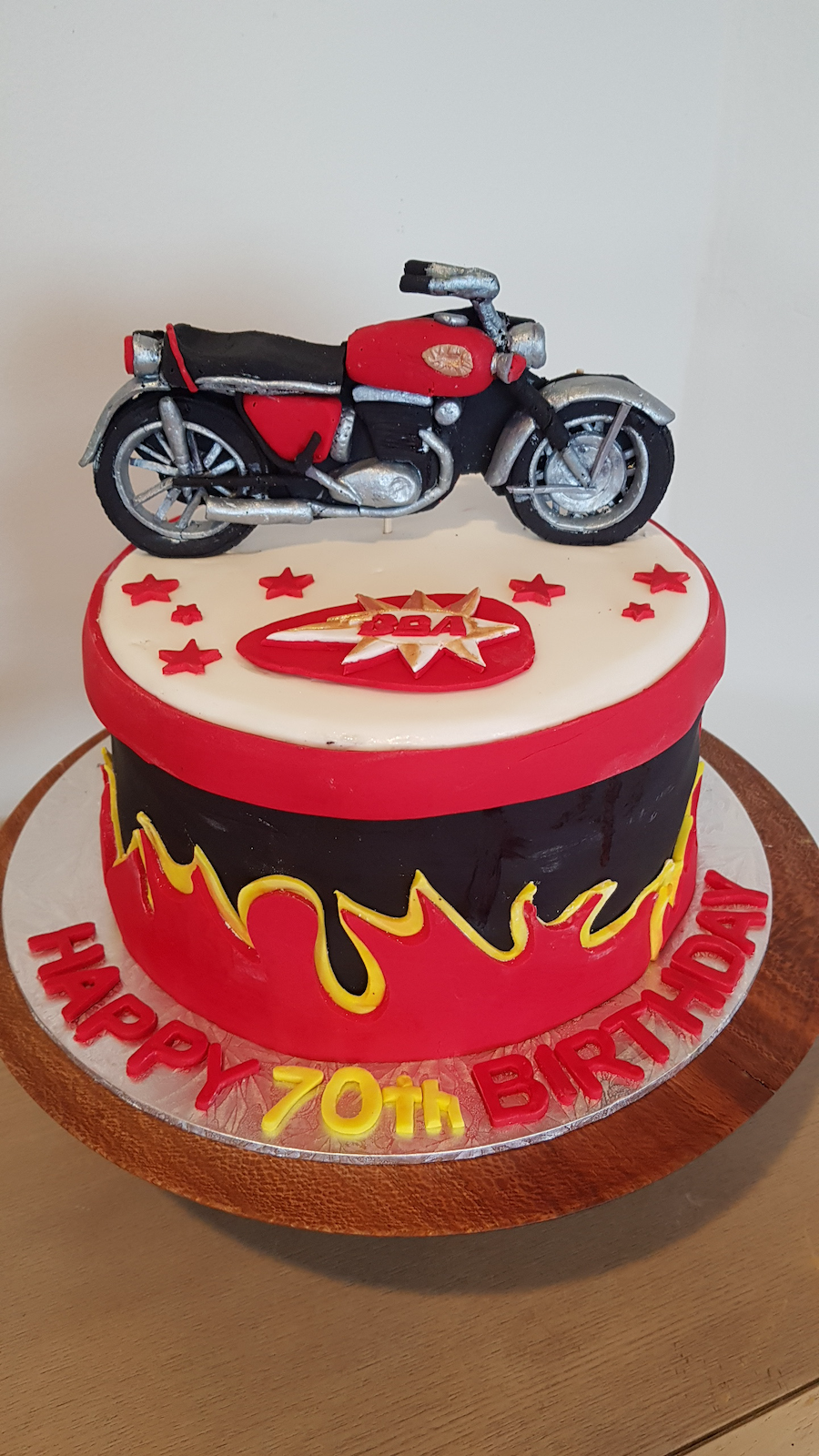 A birthday cake with a motorcycle figurine on top, decorated in red, black, and yellow flames, with the message "Happy 70th Birthday" on the base.