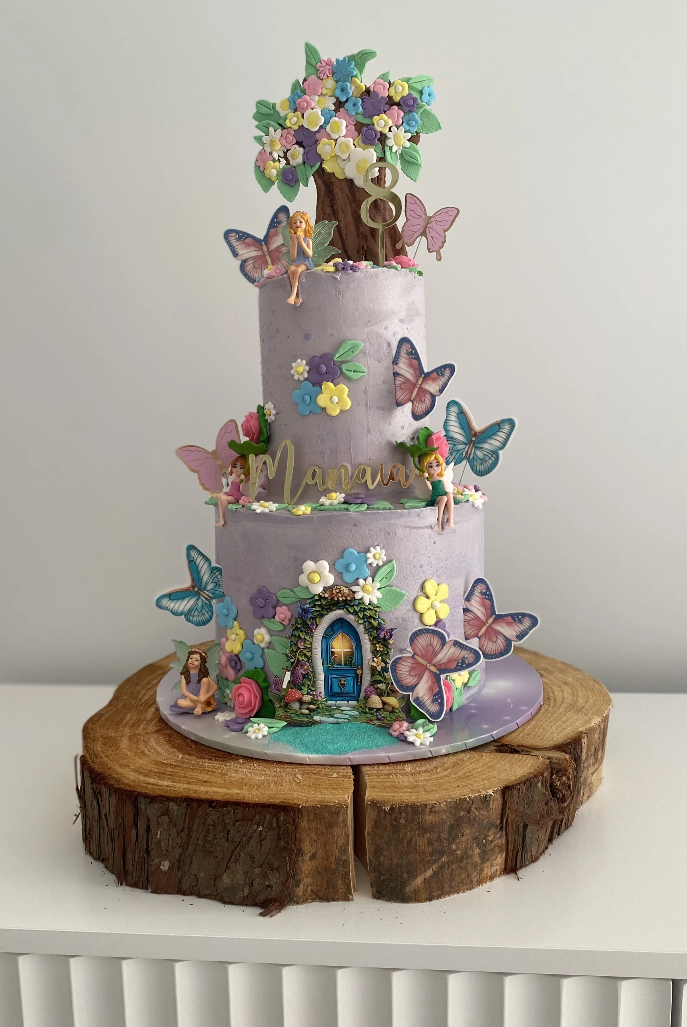 A tall, three-tier lavender-colored birthday cake decorated with colorful flowers, butterflies, fairy figures, and a wooden cutout of the name 'Manava'. The cake features a whimsical fairy tale theme, with a woodland door at the base, a tree with blo