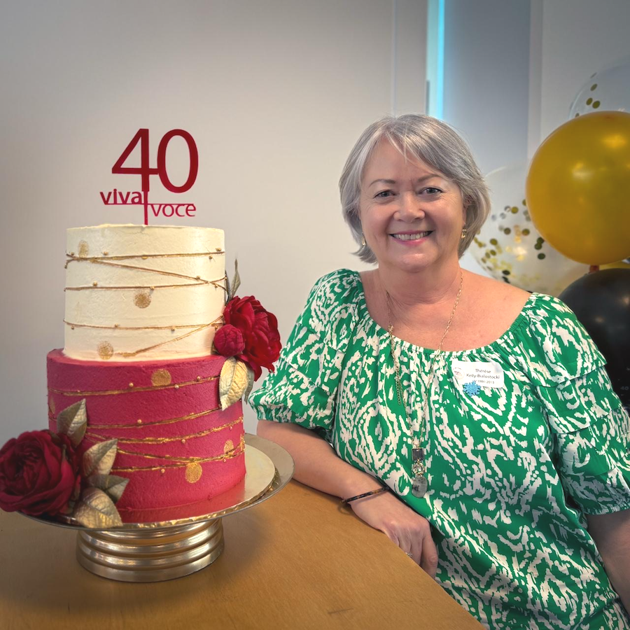 A smiling woman sitting at a table beside a decorated birthday cake with the number 40 and the words 'Viva Voce'. The cake is decorated with red and pink roses and gold details. Balloons are visible in the background.