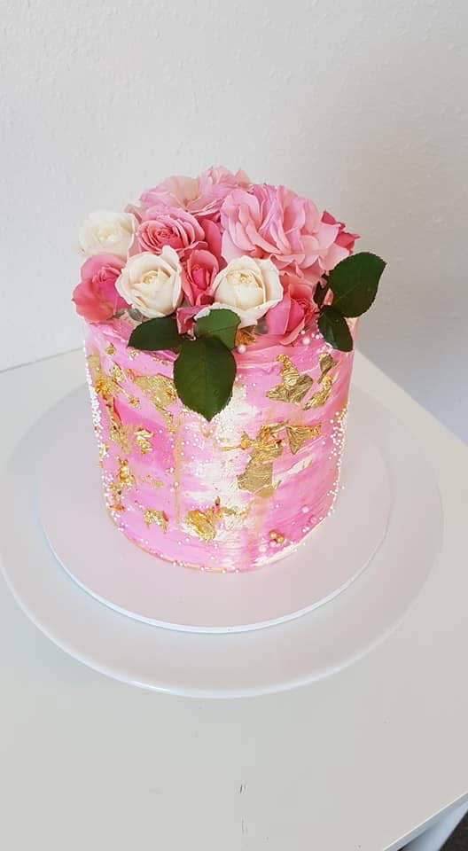 A pink cake decorated with white and pink roses, gold leaf decorations, and a few green leaves on top, placed on a white round cake board.
