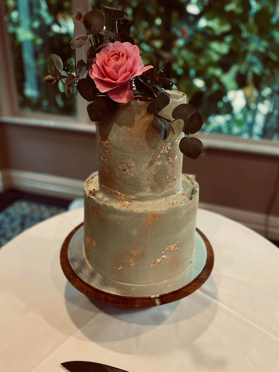 Two tiered wedding cake, lemon and coconut tier, chocolate tier, iced with white chocolate ganache coloured sage green for a 'concrete' effect with gold shimmer and gold leaf.  The fresh rose is Sam McGredy's 'My Girl'. 