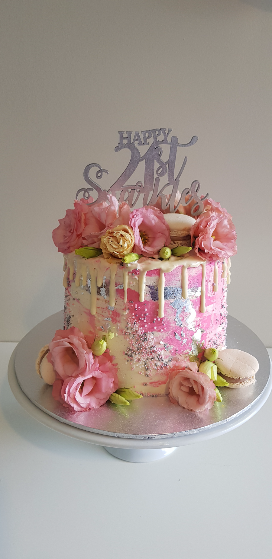 Pink and white birthday cake with floral decorations, macarons, a "Happy 21st Birthday" topper, and edible silver and colorful sprinkles.