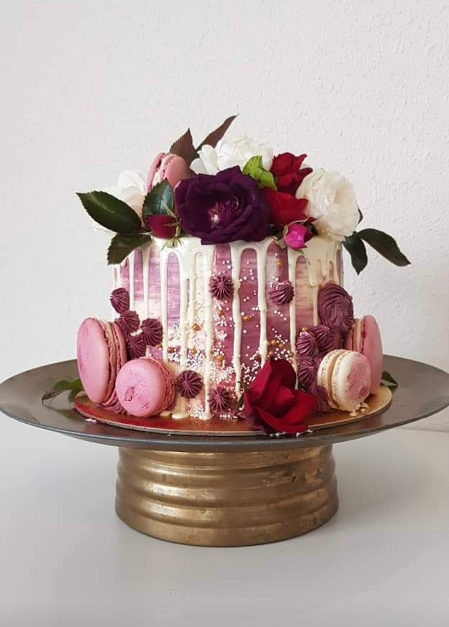 A pink and white floral decorated cake with macarons, topped with roses and floral accents, displayed on a gold pedestal