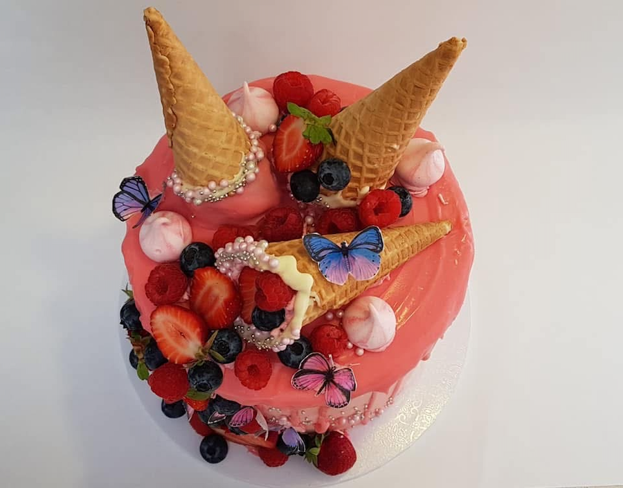 A pink heart-shaped cake topped with strawberries, blueberries, raspberries, pink and white meringues, colorful edible butterflies, pearl-like beads, and three waffle cones.