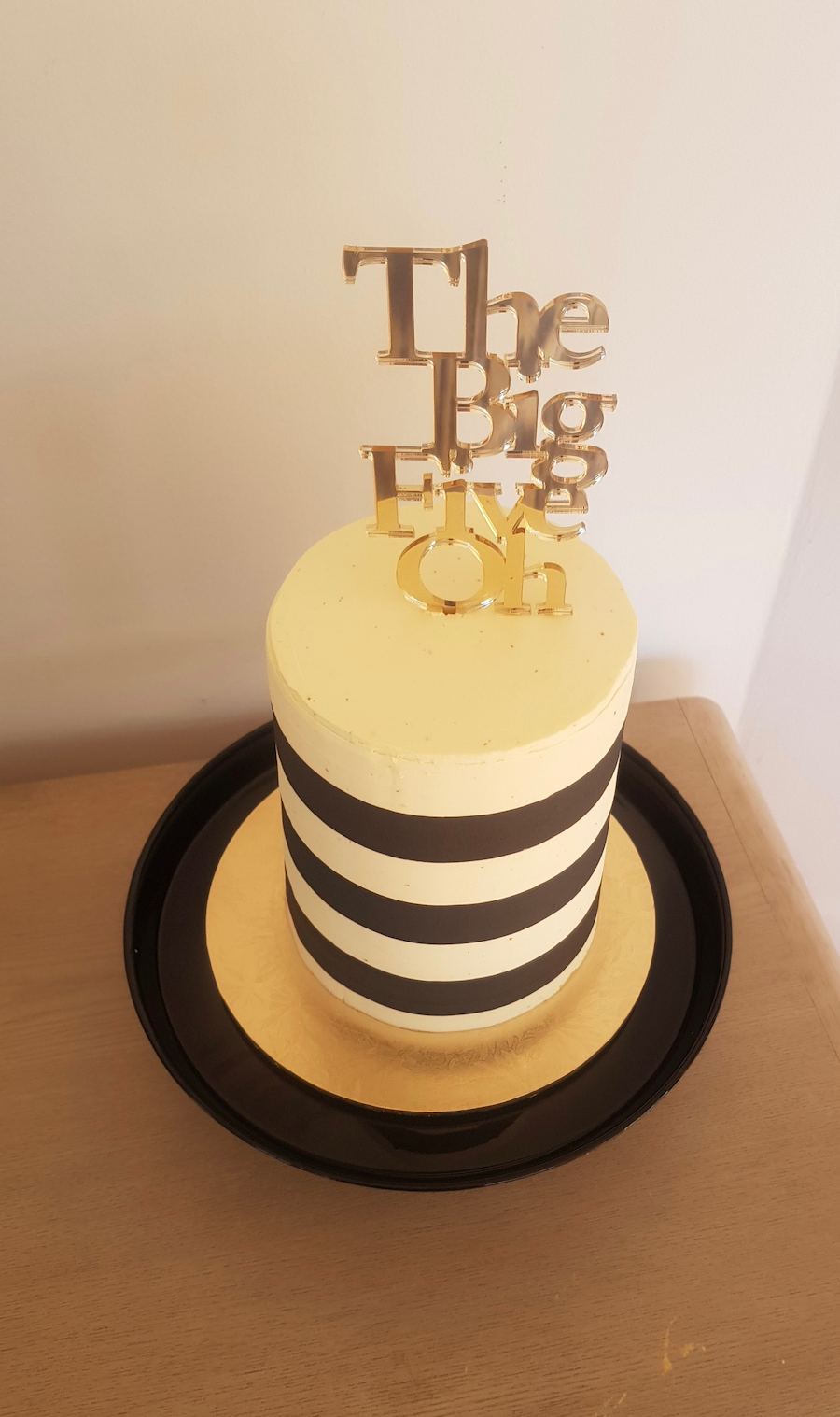 Cake with white frosting and black horizontal stripes, topped with a gold cake topper that reads 'The Big One'.