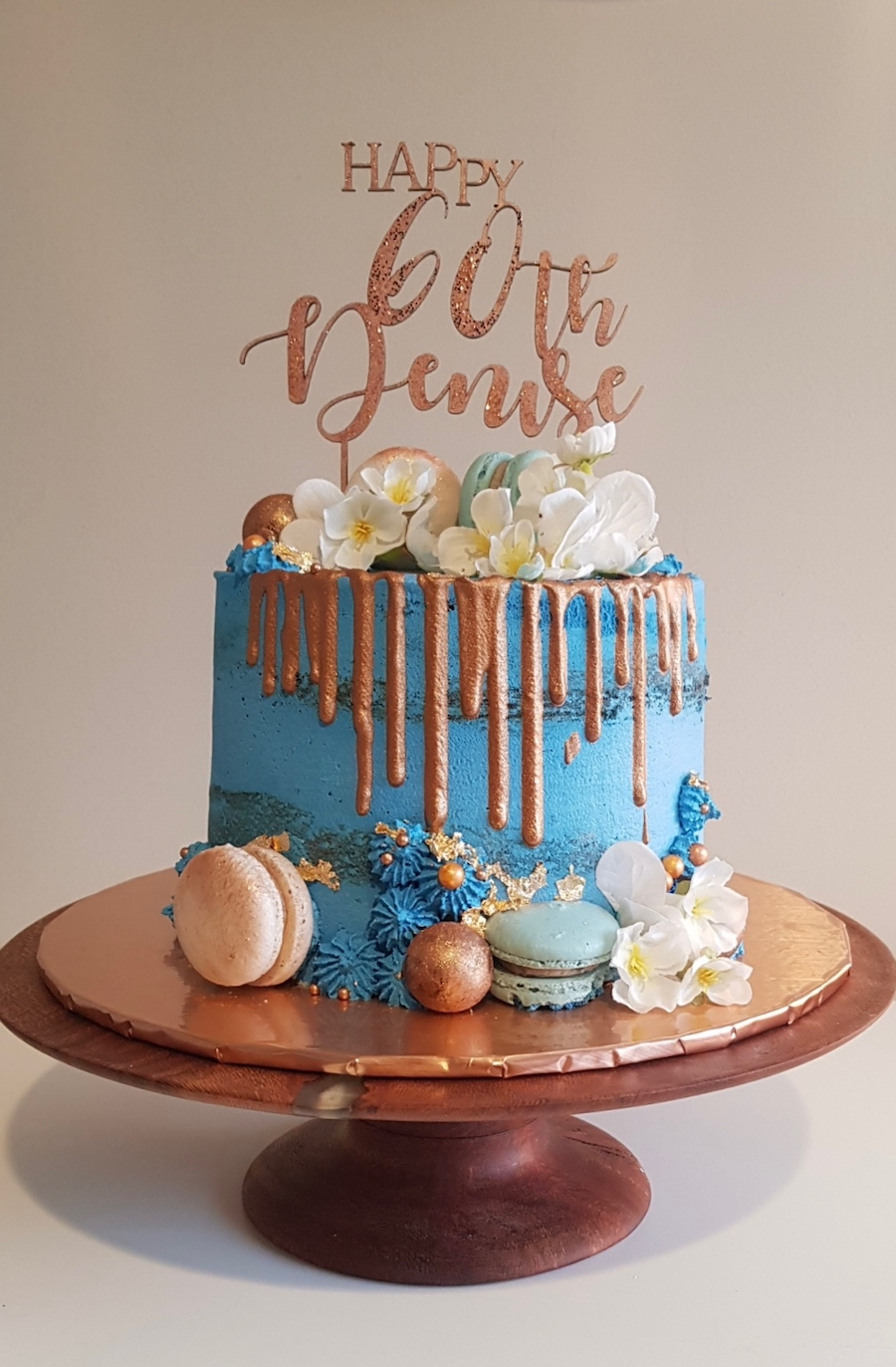 Blue birthday cake with gold drip decoration, white flowers, and macarons, with a "Happy 60th Birthday" topper on a wooden cake stand.