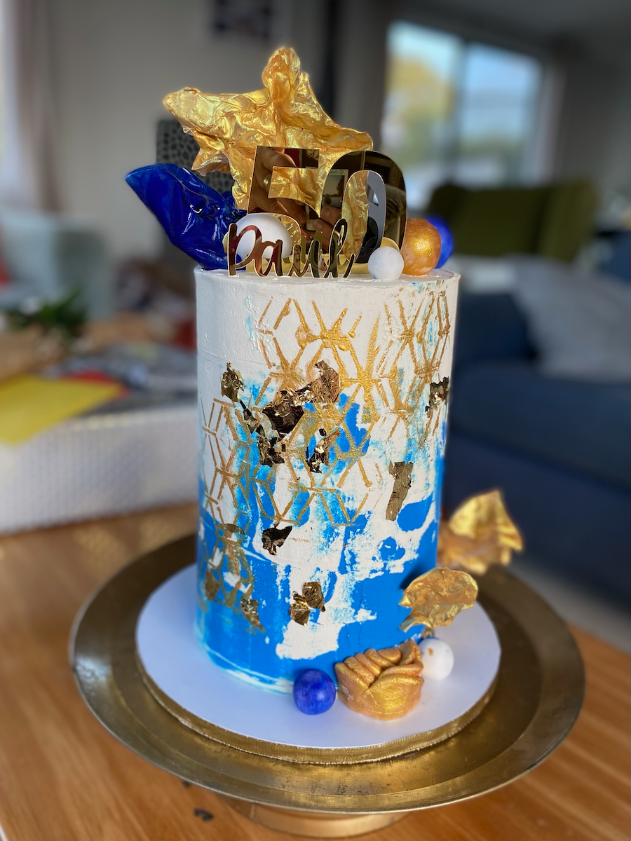 A tall birthday cake decorated with gold, blue, and white frosting, featuring gold leaf accents and geometric patterns. The cake is topped with gold, blue, and white decorative elements, including a "7" and "Paulo" with gold and silver details. The c