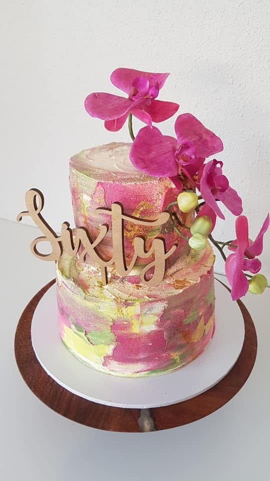 A two-tier birthday cake with pastel pink, yellow, and green watercolor-like icing, decorated with pink orchids and green berries, with a wooden word cake topper reading 'Sixty' on top.