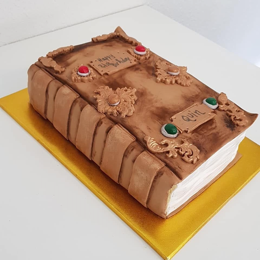 A cake decorated to look like a hardcover book with a brown cover and ornate decorations, and a small sign on top with 'Happy 21st Birthday' handwritten. The cake is placed on a gold-colored board.