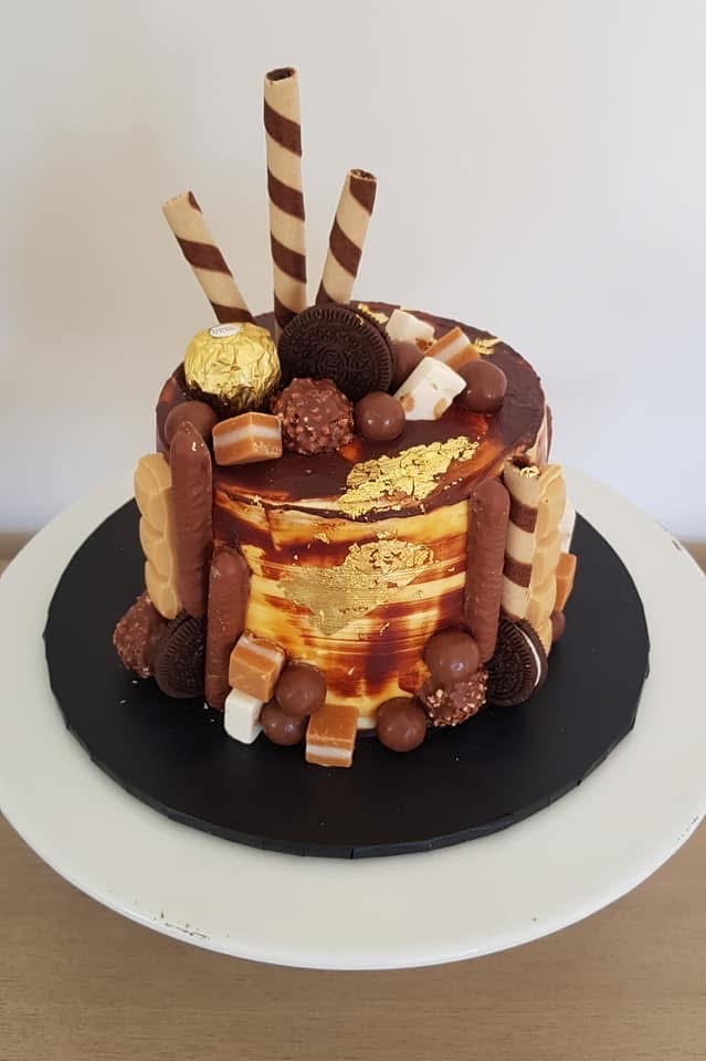 A decorated birthday cake with chocolate and caramel toppings, gold leaf accents, Oreo cookies, wafer sticks, and assorted chocolates, placed on a black and white cake board.