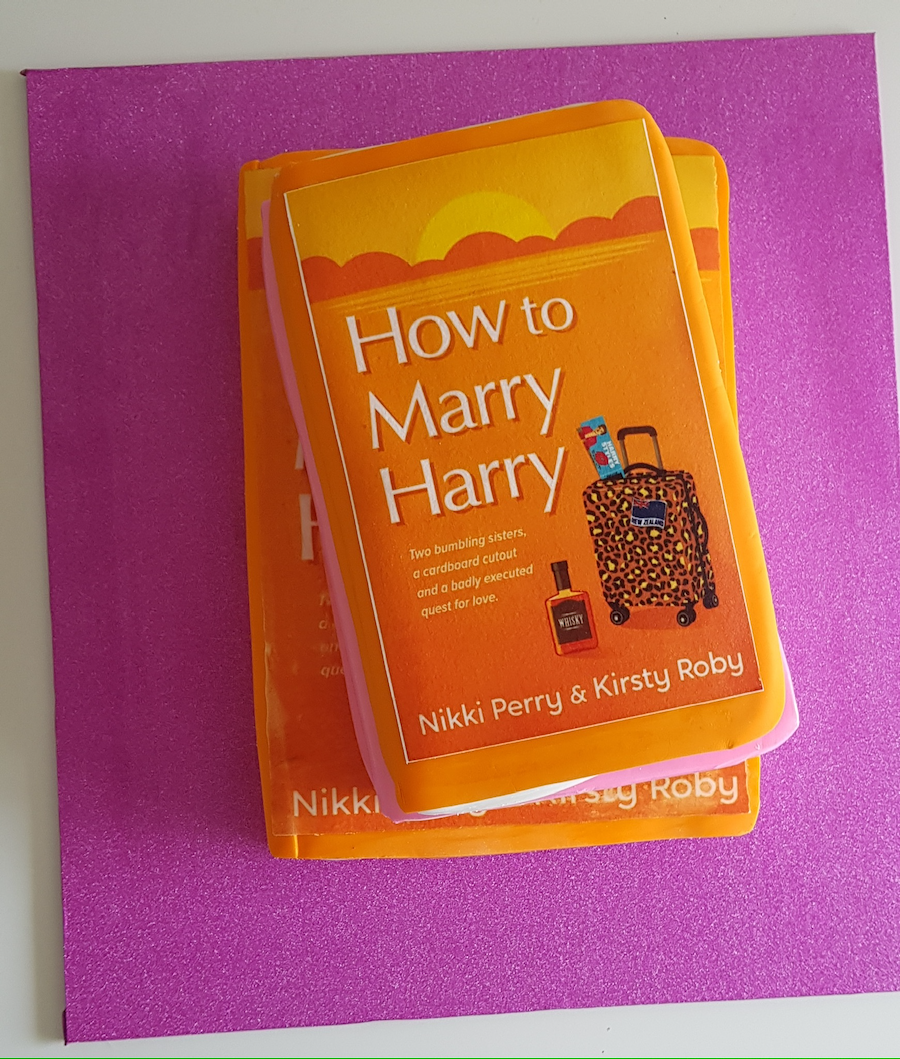 A book titled "How to Marry Harry" by Nikki Perry and Kirsty Roby, with a subtitle about two sisters on a quest for love, placed on a pink surface.