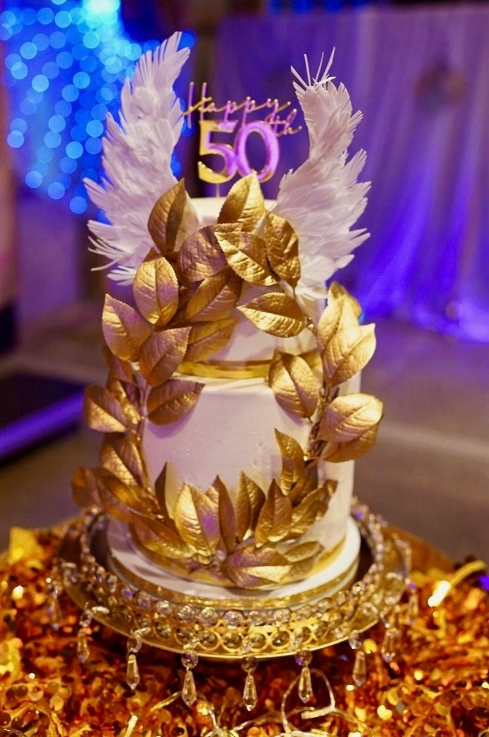 A white birthday cake celebrating a 50th birthday with gold leaves, white feather decorations, and a gold topper that says 'Happy 50th'. The cake is placed on a gold ornate stand with hanging jewel embellishments, surrounded by gold confetti.