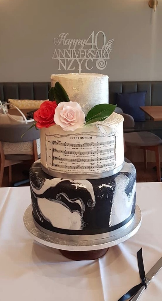 A three-tiered cake with black and white marbled design on the bottom tier, sheet music and floral decorations on the middle tier, and a plain cream top tier. The cake is decorated with real flowers and a topper that reads "Happy 40th Anniversary NYC