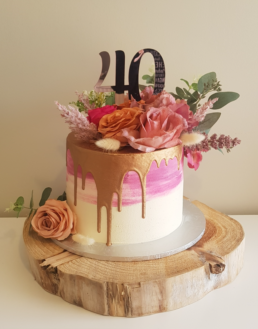 Decorative cake with pink and gold drip, topped with pink and purple flowers, green foliage, and a "40" cake topper on a wooden slab.