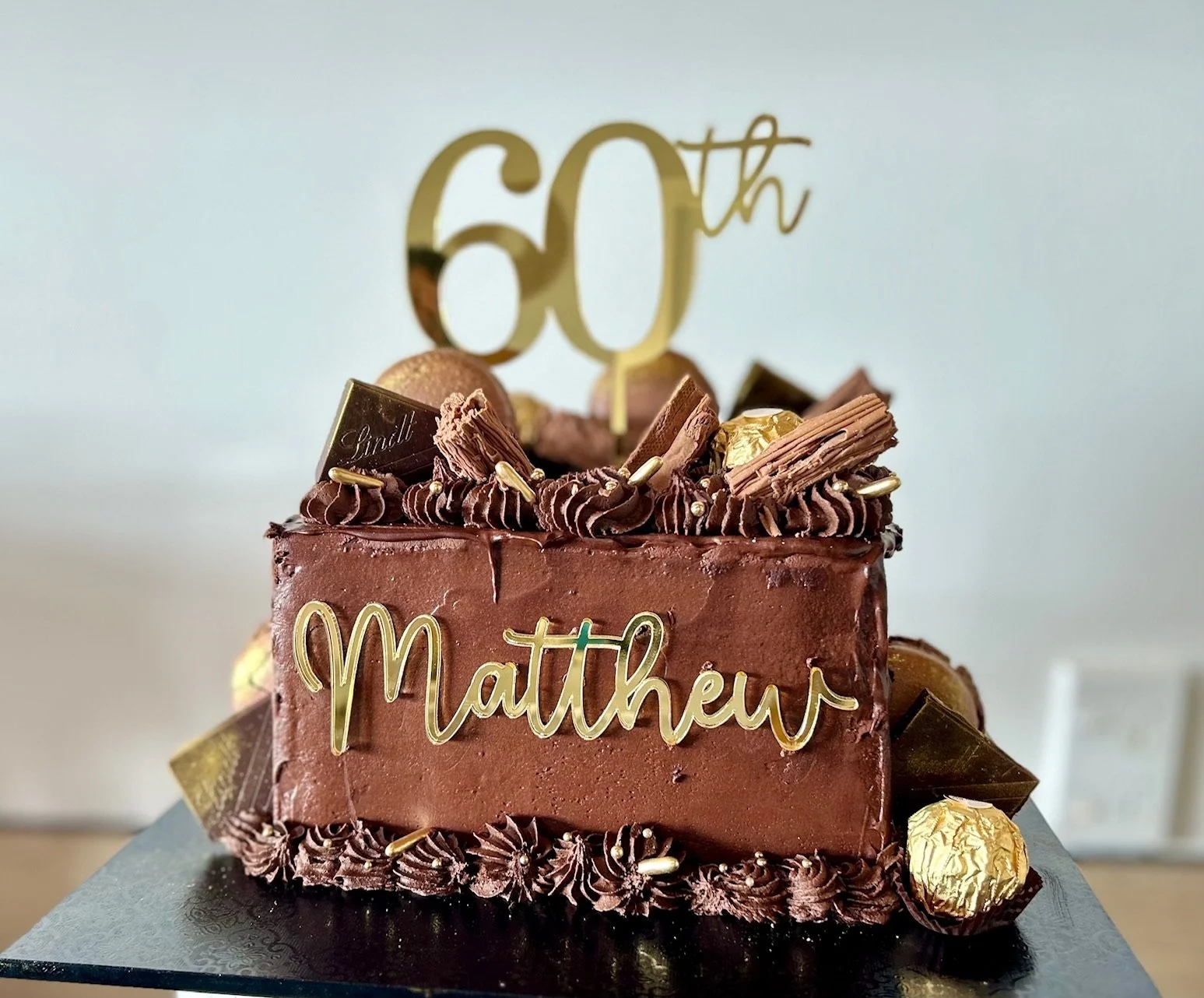 A square carrot cake with cream cheese filling and chocolate ganache frosting, gold accents and topper.