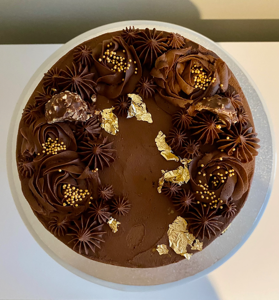 Chocolate cake decorated with chocolate frosting rosettes, gold and chocolate sprinkles, chocolate-covered treats, and gold leaf accents, on a white cake stand.