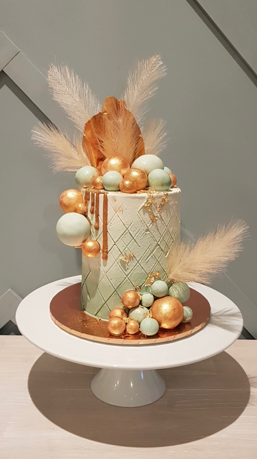 A tall, cylindrical cake decorated with gold and pastel green spherical accents, gold drips, and fluffy pampas grass on a white cake stand.