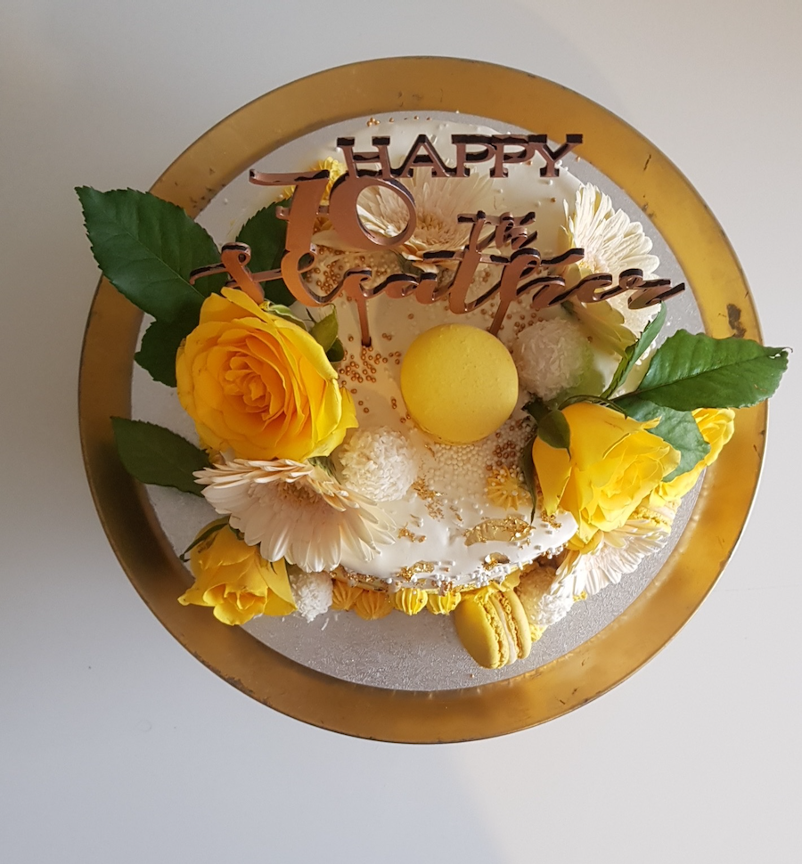 Celebratory cake with yellow roses, white flowers, macarons, and a "Happy 70th Birthday" topper.