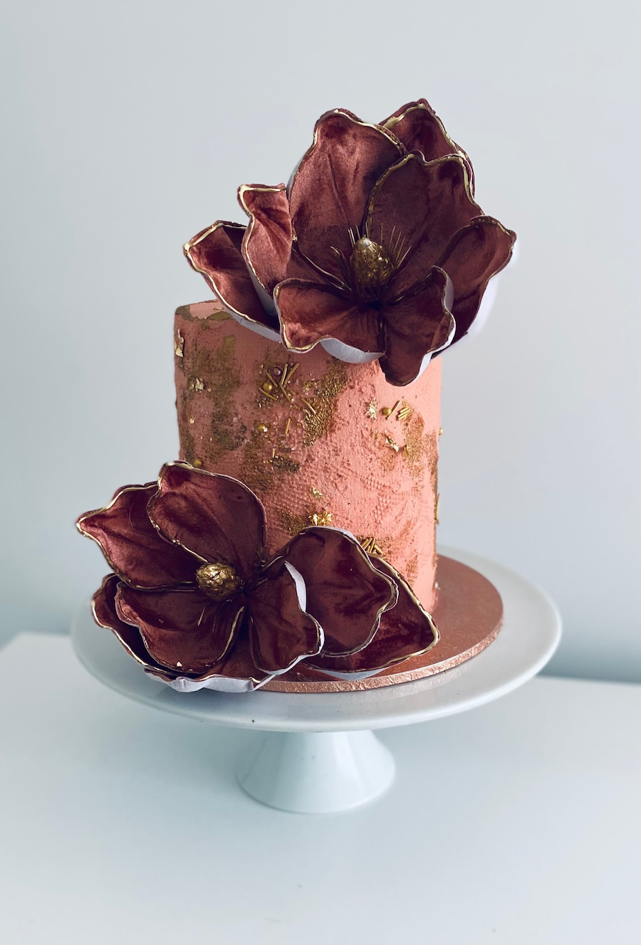 A pink, textured cake decorated with large, dark red flowers with gold accents, placed on a white cake stand against a plain background.