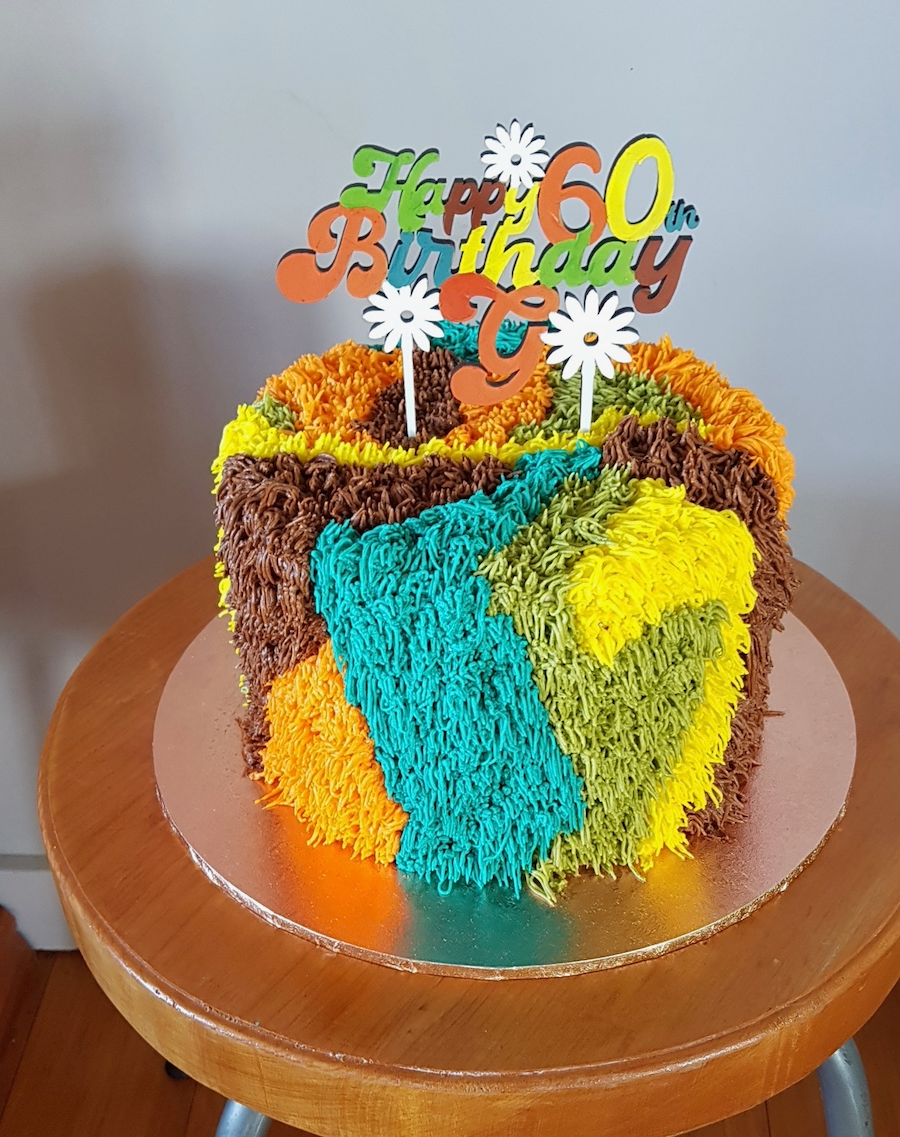 Colorful birthday cake with a brown, yellow, blue, green, and orange icing design, placed on a wooden table, with a "Happy 60th Birthday" cake topper.